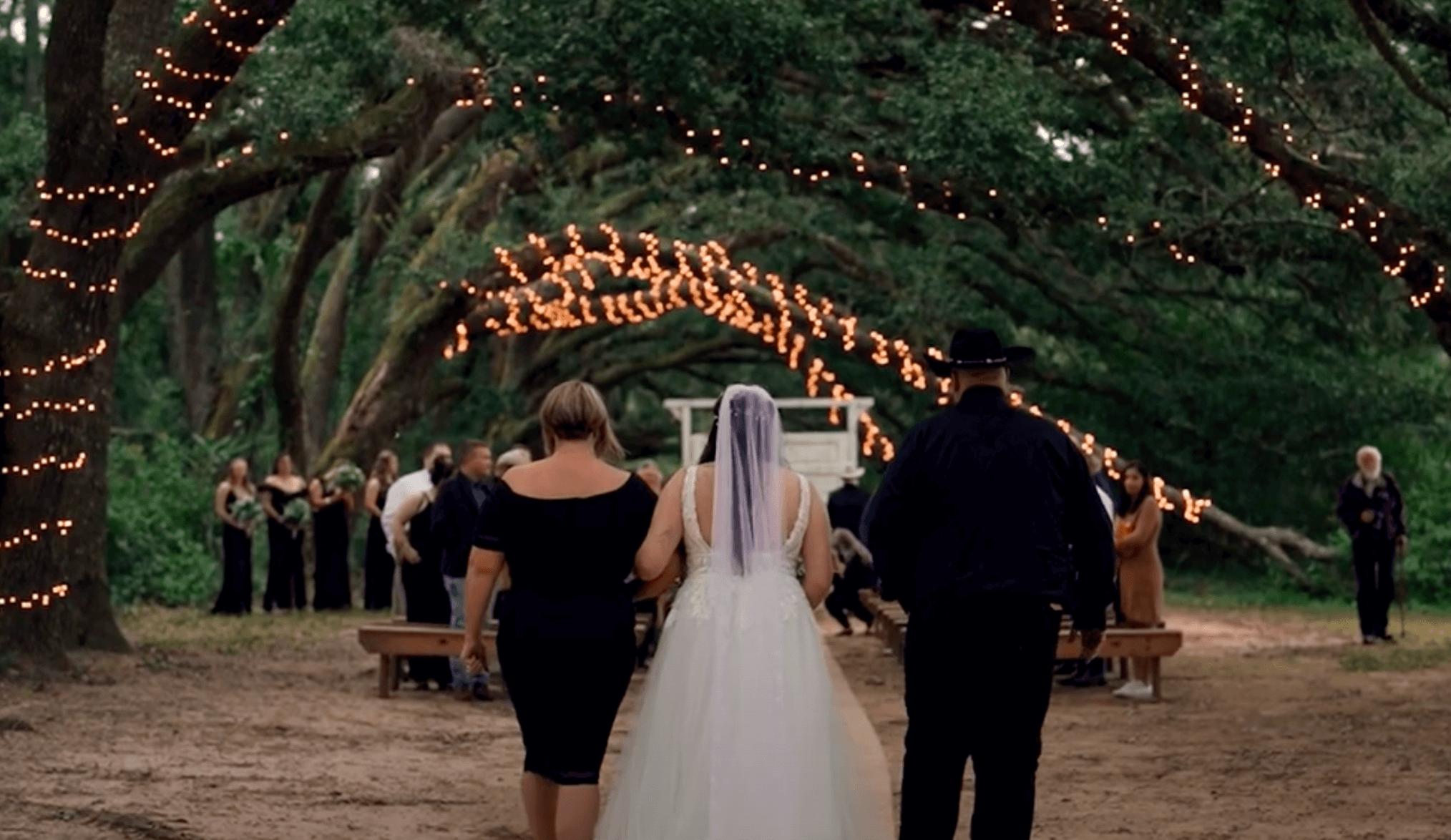 Bride walking down the aisle outside under a canopy of lit oak trees with her mom and dad