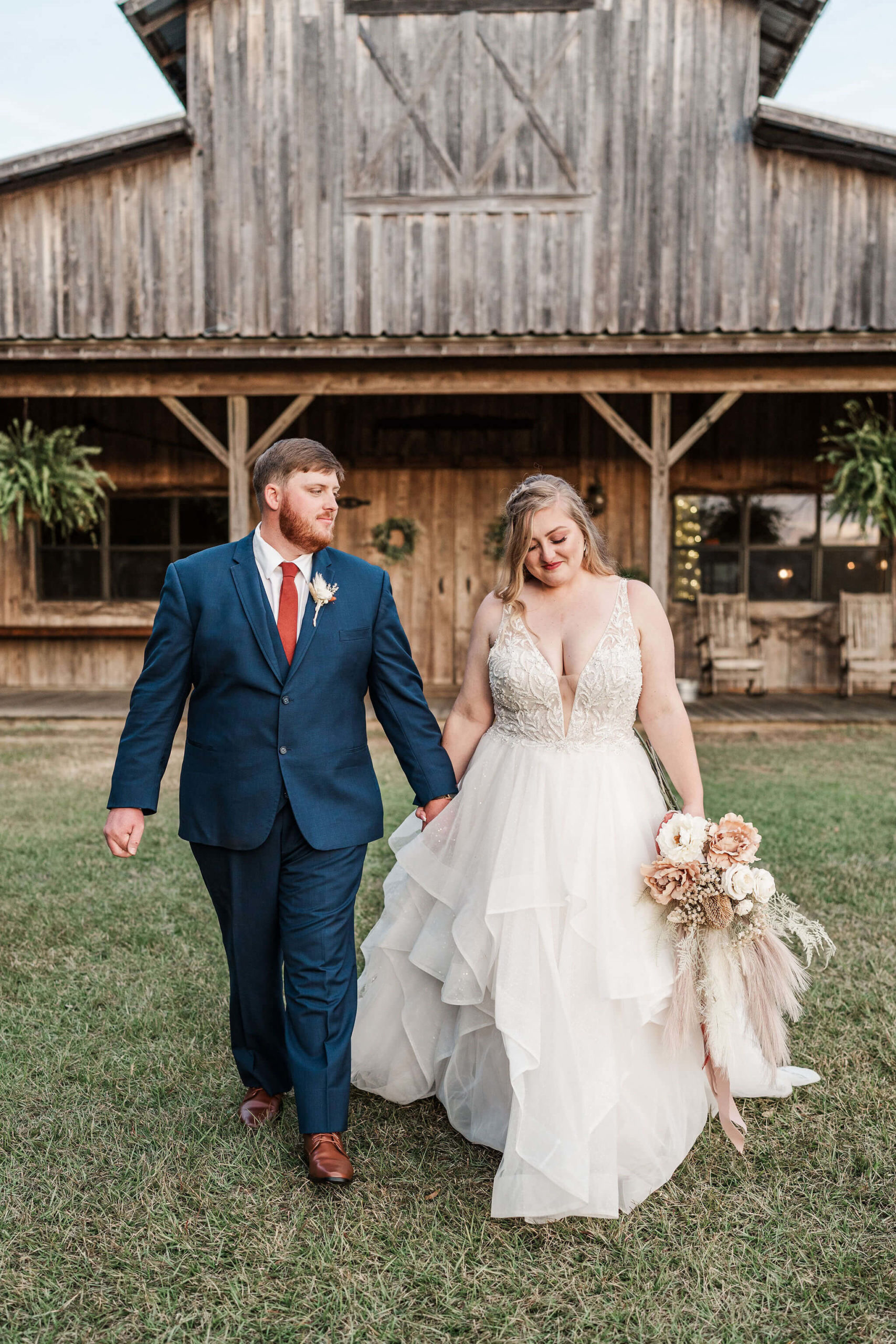 Bride and groom walking in front of rustic barn. Groom is smiling at bride while she's looking down at her dress