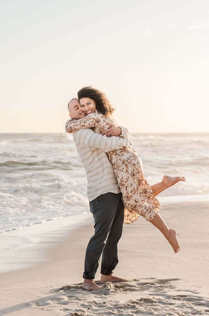 Man picks up his and wife-to-be while she kicks up a leg and smiles for their Orange Beach Alabama Point engagement photos