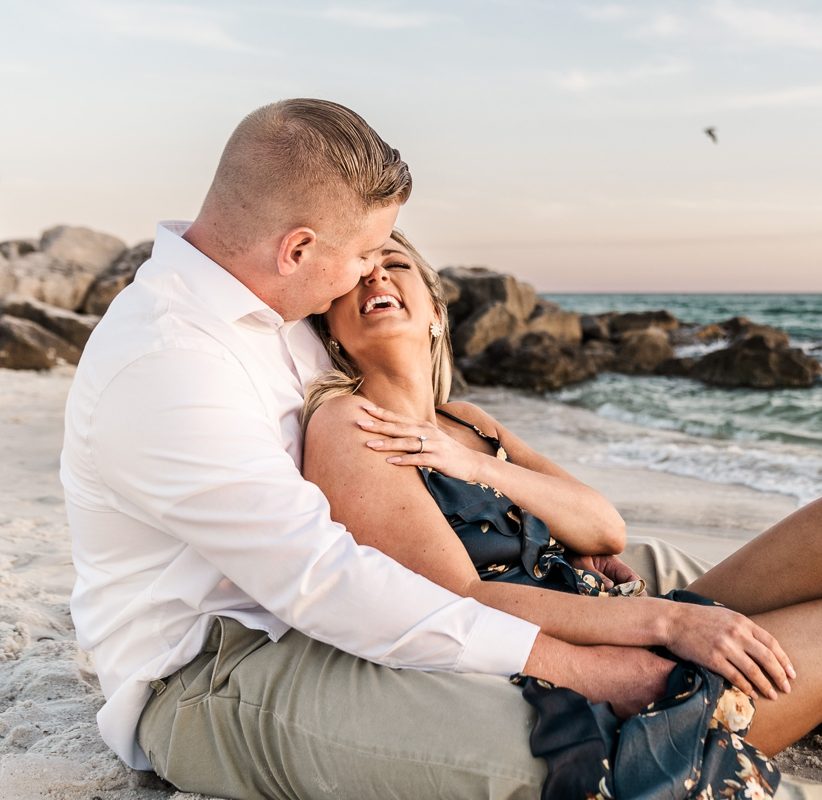 Man and woman sit down on beach while laughing for their Alabama Point engagement photos.