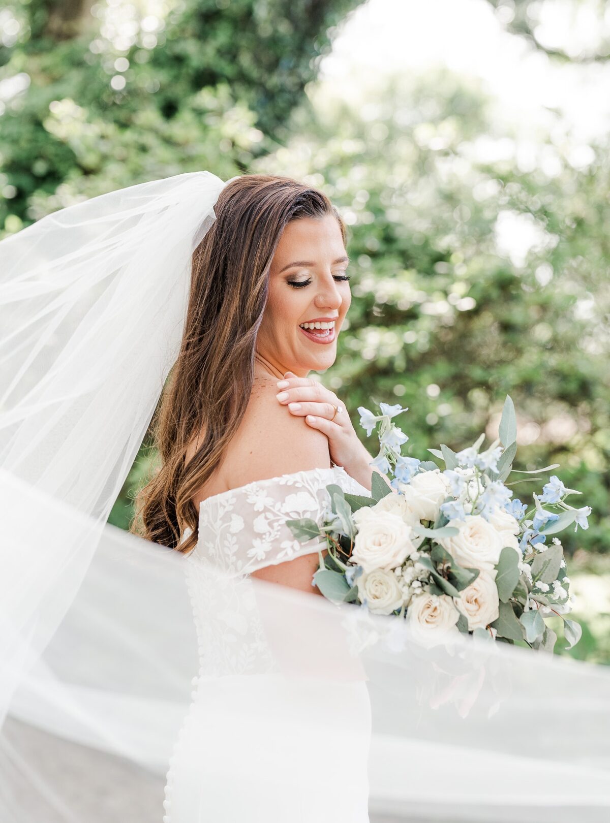 Bride holds bouquet and smiles down her right shoulder as the wind takes her veil in the air for her Bragg-Mitchell Mansion bridal photos.