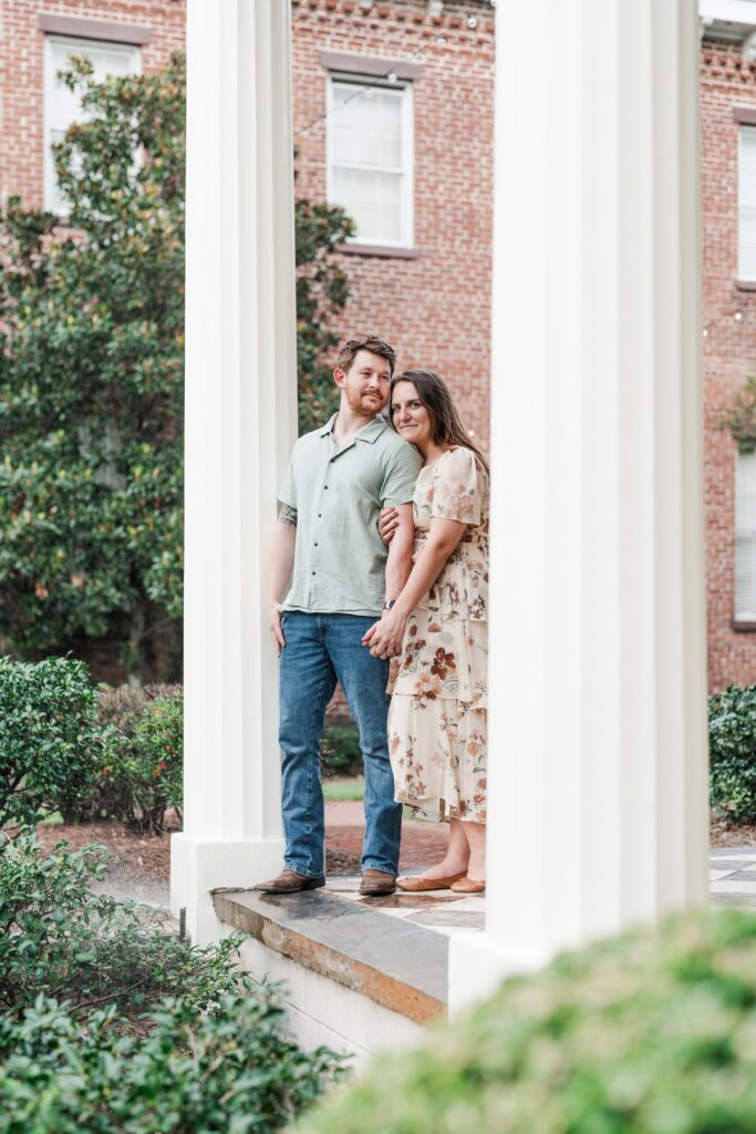 Man leans on white column while looking off in the distance and woman snuggles in behind  and smiles at the camera for their Fort Conde Inn engagement photos in Mobile, Alabama
