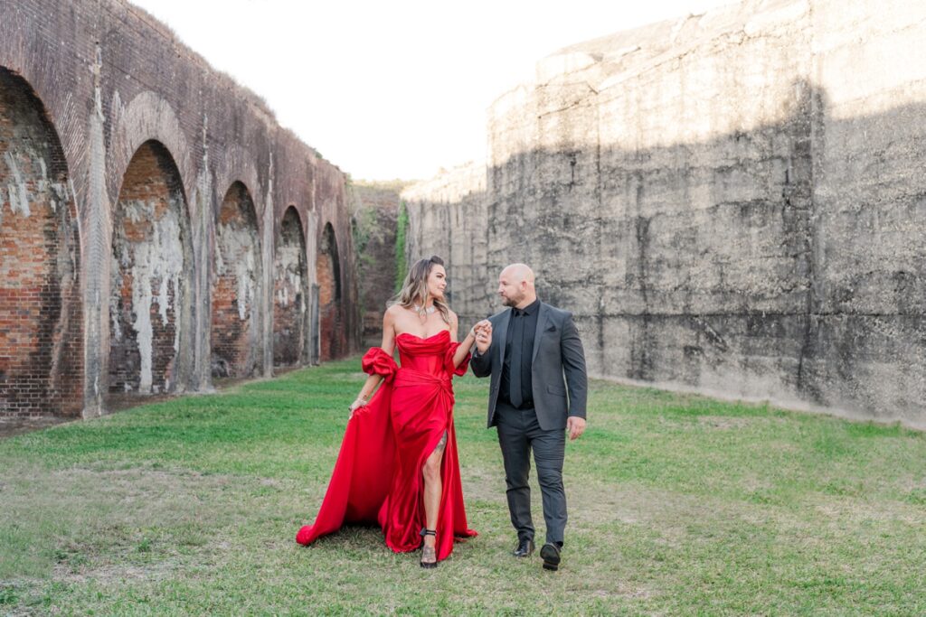 man in black suit and woman in red silky dress walk in abandoned historic fort battleground site